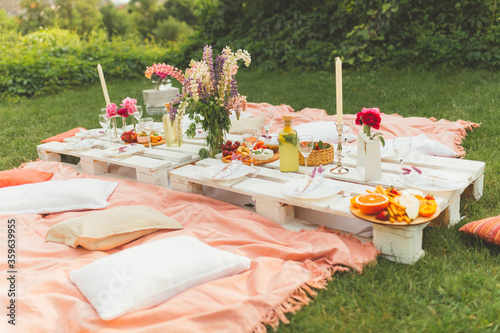 Celebration summer picnic in the city garden. Delicious food and drinks on pallet tables.  Decorated with beautiful flowers and candles. Rest and leisure for family and friends. Pleasure time