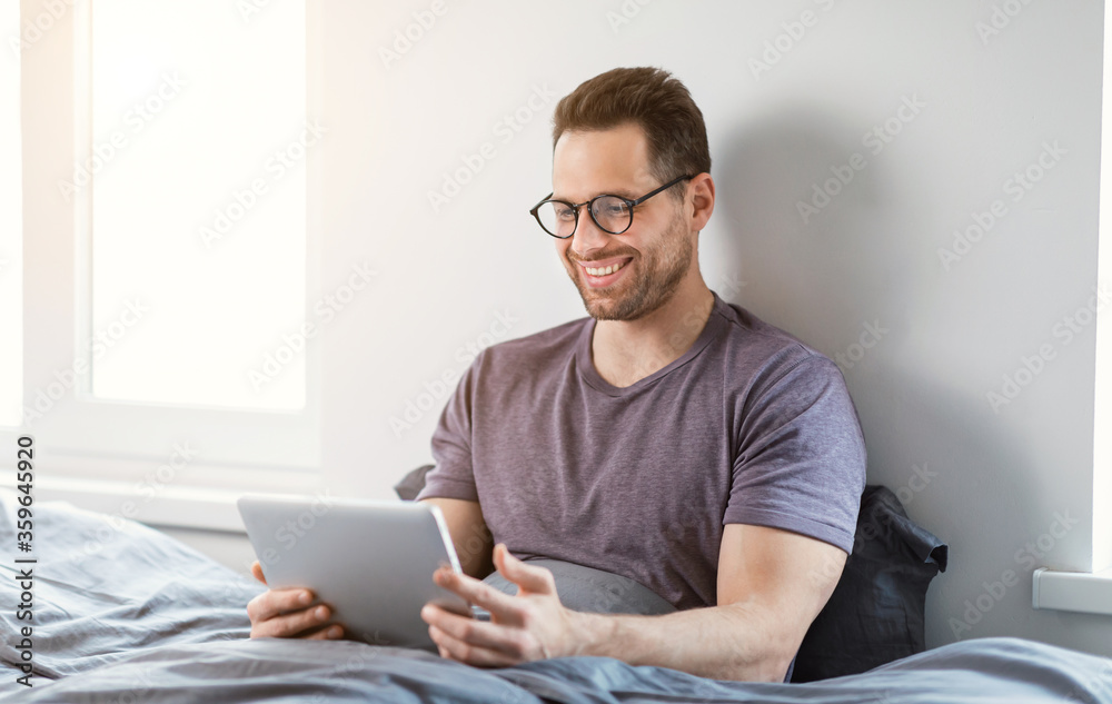 Happy Man Using Tablet Browsing Internet Sitting In Bed Indoor