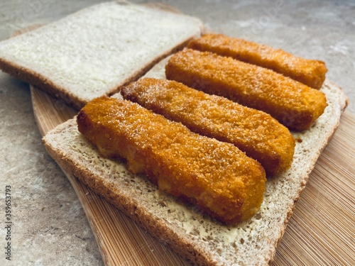 Closeup view of crispy fish fingers covered in salt and vinegar on buttered bread in the kitchen. Breaded fish finger sandwich preparation on wooden board.