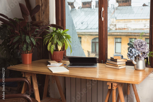 Wooden working desc tabletop with green indoor plants, laptop, books, and notebooks. Window view, rainy autumn weather