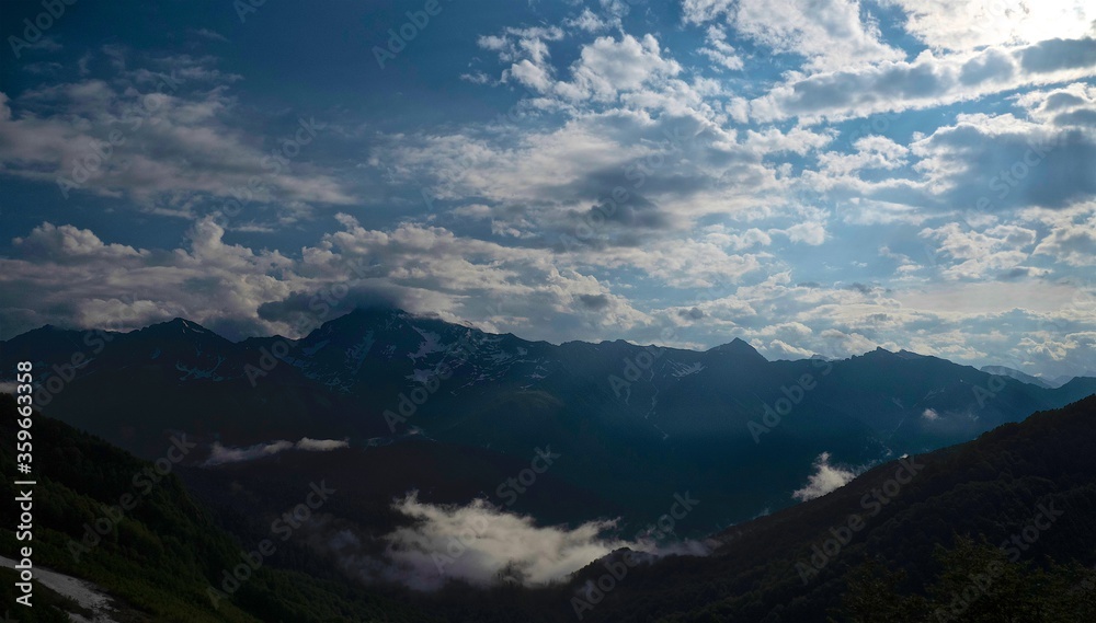 Morning fog creeps along the valley among the high mountains. Early morning in the mountains. The tops of pines stick out of the fog. Morning after a night shower. Blue sky with light clouds. Morning fog creeps along the valley among the high mountains. Early morning in the mountains. The tops of pines stick out of the fog. Morning after a night shower. Blue sky with light clouds.
