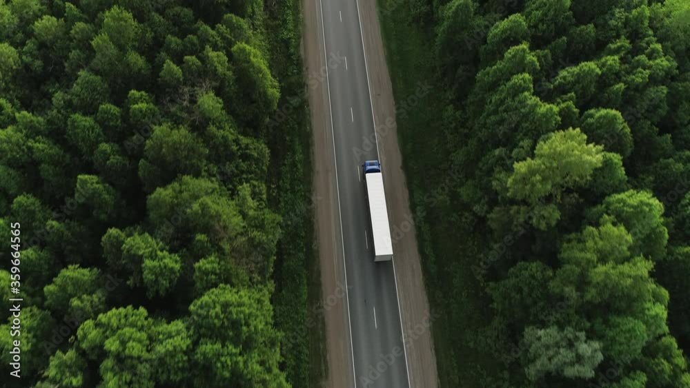 One Semi Truck with white trailer and blue cab driving / traveling alone on dense flat forest asphalt straight empty road, highway top down view follow vehicle aerial footage / Freeway trucks traffic One Semi Truck with white trailer and blue cab driving / traveling alone on dense flat forest asphalt straight empty road, highway top down view follow vehicle aerial footage / Freeway trucks traffic