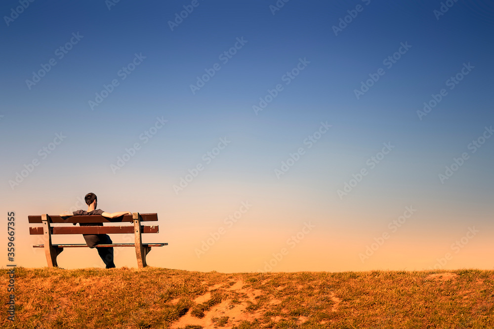 young man resting alone on a bench in an empty landscape at dusk - obrazy, fototapety, plakaty 