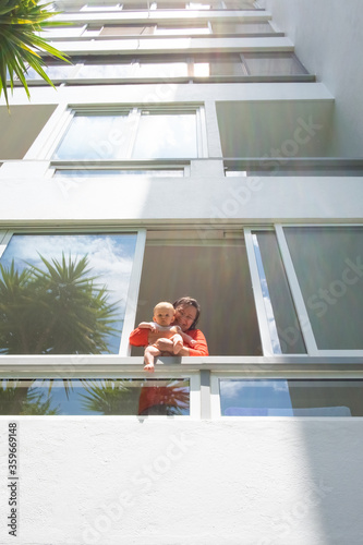 Happy grandma holding baby and leaning on balcony railing, standing inside apartment, sticking out from open window. Outdoor shot, facade view. Child care or staying at home concept