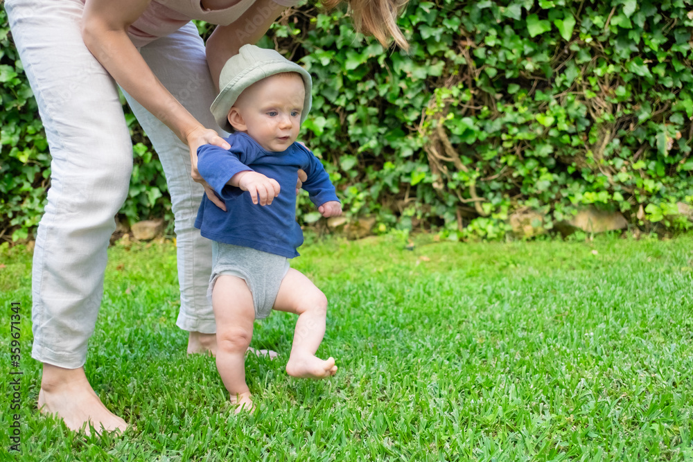 Lovely baby girl in hat and blue shirt doing first steps with help of mother. Young pretty mom holding daughter on grass. First barefoot steps, garden and sunny days concept Lovely baby girl in hat and blue shirt doing first steps with help of mother. Young pretty mom holding daughter on grass. First barefoot steps, garden and sunny days concept