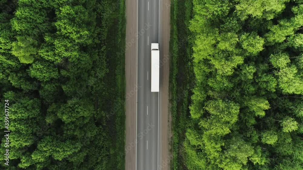 One Semi Truck with white trailer and cab driving / traveling alone on dense flat forest asphalt straight empty road, highway top down view follow vehicle aerial footage / Freeway trucks traffic