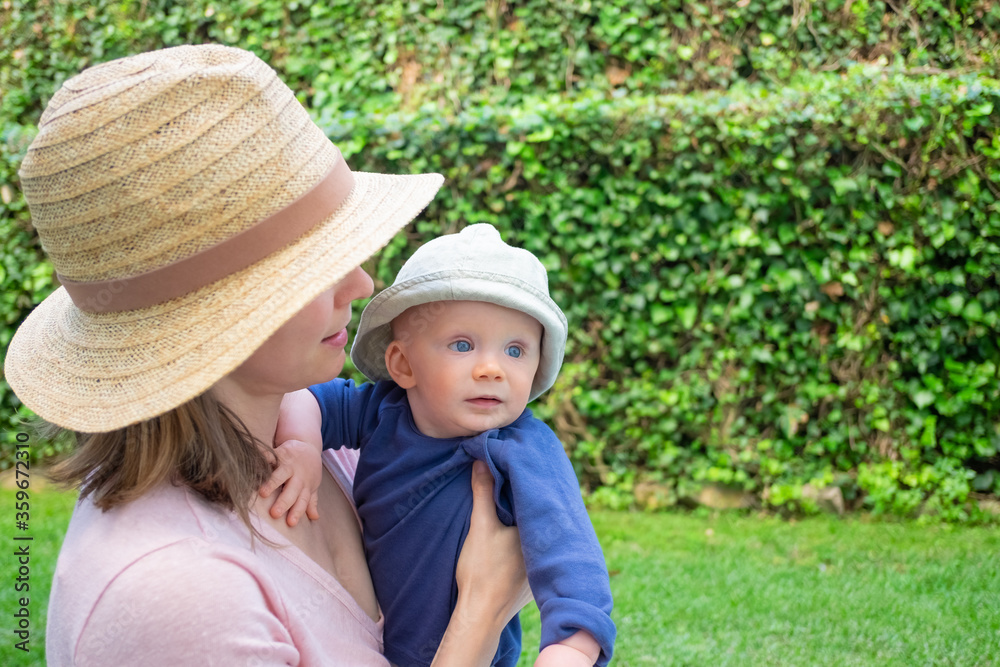 Young mother in hat holding daughter in park and looking at her. Adorable baby girl in hat with blue eyes looking away. Summer family time, garden and sunny days concept
