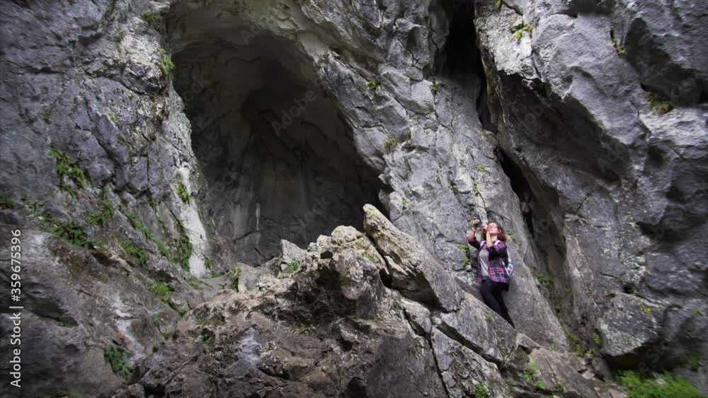View from below of young woman taking photo of mountains standing on high cliff near cave hole. Russia View from below of young woman taking photo of mountains standing on high cliff near cave hole. Russia