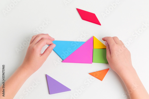 Tangram colored geometric puzzle pieces with childs hands moving pieces on white table, top view. 