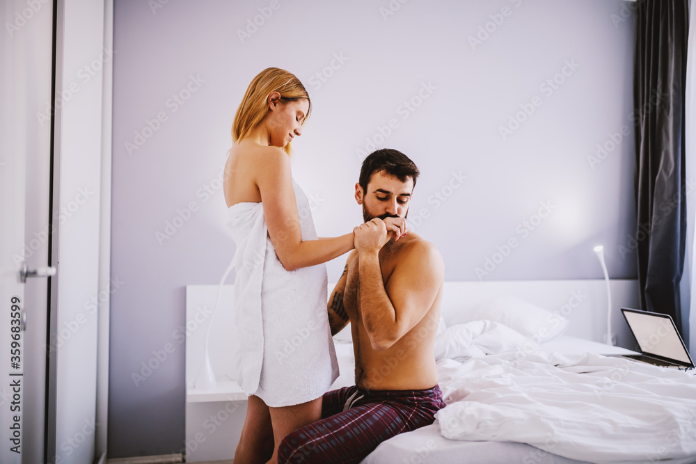 Young woman wrapped in towel standing and seducing her boyfriend. Man sitting on bed and kissing her hand. Young woman wrapped in towel standing and seducing her boyfriend. Man sitting on bed and kissing her hand.