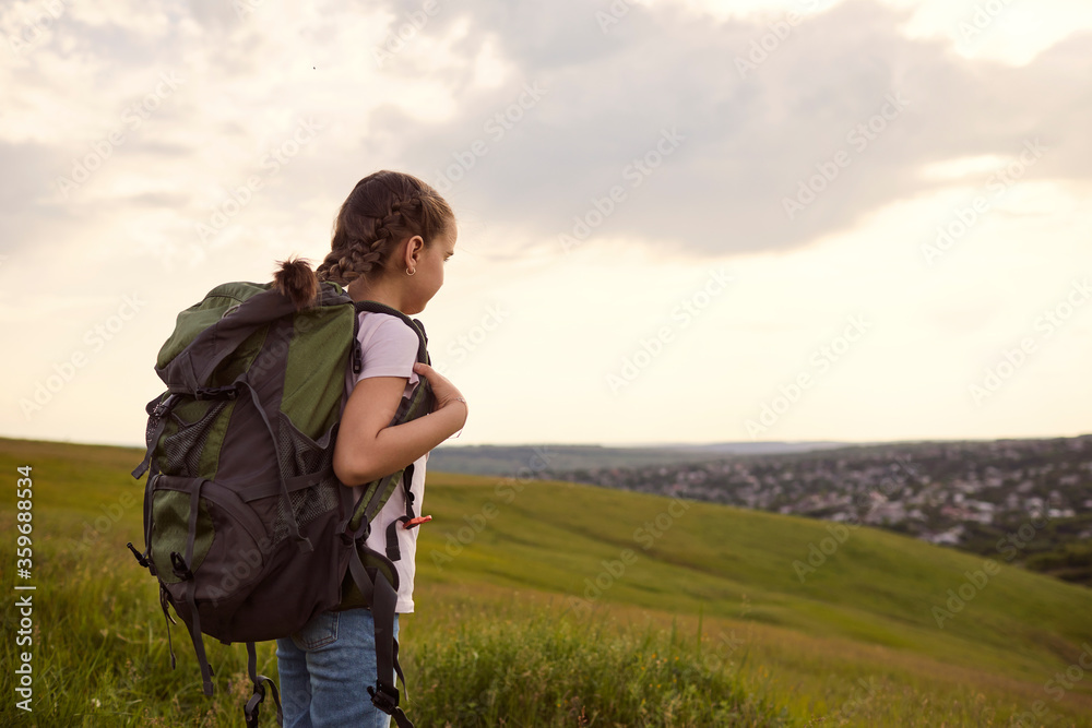 Little girl tourist with a backpack on a hiking strolls forward in nature back view.