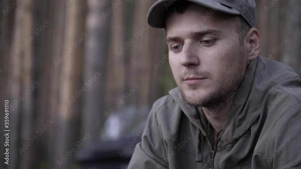Portrait of young adult male tourist sits on log near fire in forest. Human looks at forest fire on nature. Cooking dinner on wild hike. Outdoor travel. Close up shot
