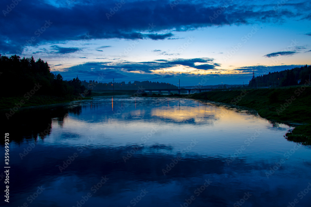 Silhouettes of trees and a bridge under a dark sunset sky that is reflected in the river.