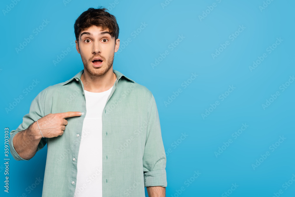 shocked young man pointing with finger while looking at camera on blue shocked young man pointing with finger while looking at camera on blue