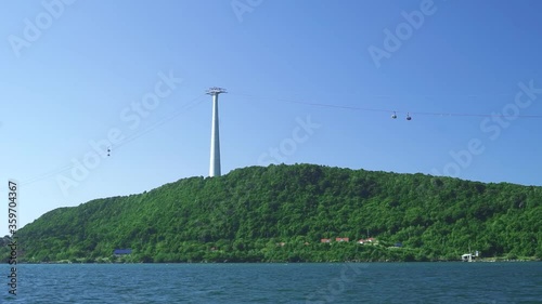 A view of a green island in the bue sea with aerial tramway pylon and moving cable cars. View from the ferry boat window in the ocean, Cable cars ascending through the aerial ropeway.