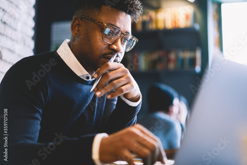 Skilled afro american IT developer in glasses concentrated while coding app for outsourcing cooperating online with testers using laptop computer and free wireless connection in coworking space