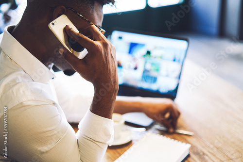 Busy afro american male entrepreneur having important phone conversation discussing ideas of new startup while sitting in cafe using laptop computer with mock up screen connected to 5G wireless