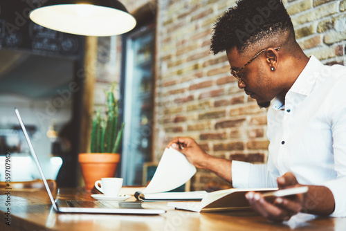 Concentrated afro american male finance advisor checking documentation and accounting reports of company income to create successful strategy for increasing sales working in cafe using gadgets