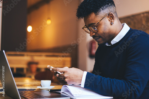 Skilled copywriter in style spectacles typing on smartphone message with phone number of cool designer and sending it to colleague for working on new project while reading books and working at laptop