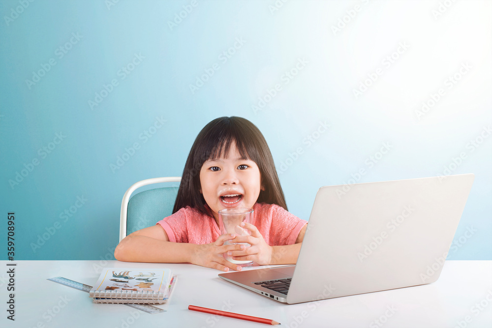 Portrait of little Asian girl drinking milk while using laptop, sitting at desk and doing homework isolated on blue background. E-learning and education concept. Portrait of little Asian girl drinking milk while using laptop, sitting at desk and doing homework isolated on blue background. E-learning and education concept.