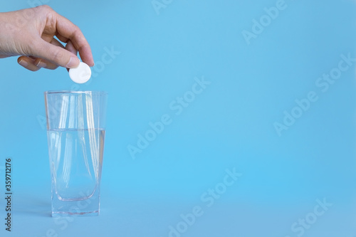 hand holds a white pill in his hands over a glass of water. Blue background.