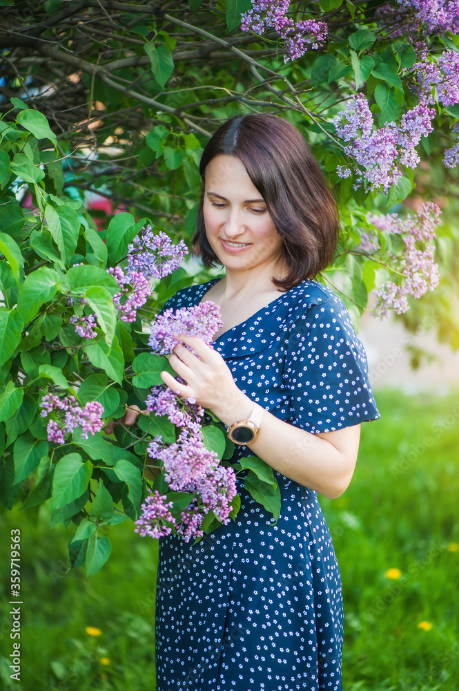 A cute brunette girl in a blue dress stands in a park, touches lilac flowers and looks at them. Soft light, vertical photo. A cute brunette girl in a blue dress stands in a park, touches lilac flowers and looks at them. Soft light, vertical photo.