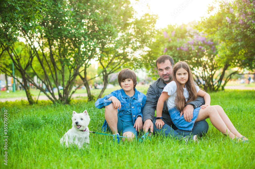 Dad with daughter and son are sitting on the grass in the park, daughter is on the mans knees. A boy in shorts and a denim jacket. In the background are trees. Nearby is a cute white dog.