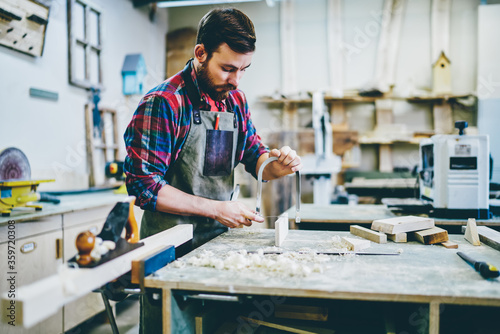 Serious bearded male joiner making details by himself using sharp tools for timber on working space, concentrated foreman in apron repairing wooden staff with instruments in workshop with sawdust. photo