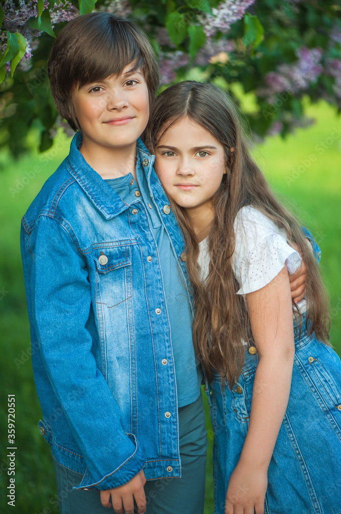 The younger pretty sister leaned on her older brother, a portrait of a girl and a teenager against a background of green grass and lilacs. Beautiful children. Soft focus, close up, vertical photo. The younger pretty sister leaned on her older brother, a portrait of a girl and a teenager against a background of green grass and lilacs. Beautiful children. Soft focus, close up, vertical photo.