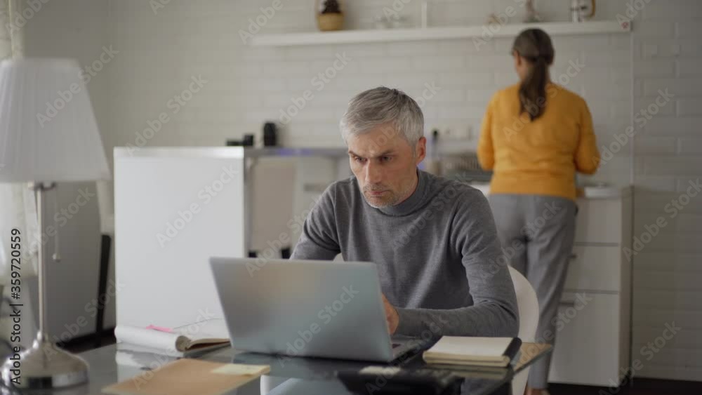 Tracking medium shot of senior man working on laptop computer and thinking sitting at table at home while his wife washing dishes in kitchen Tracking medium shot of senior man working on laptop computer and thinking sitting at table at home while his wife washing dishes in kitchen