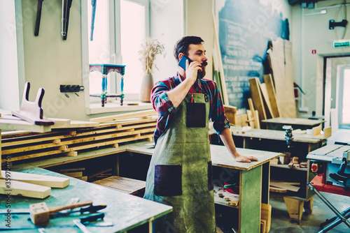 Caucasian craftsman in apron making important cellphone conversation for discussing delivery time of wooden construction to own self employed manufacturing factory, concept of online communication