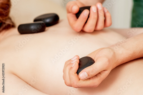 Male hand holding large new oval black hot stones for back massage on female back is spa salon