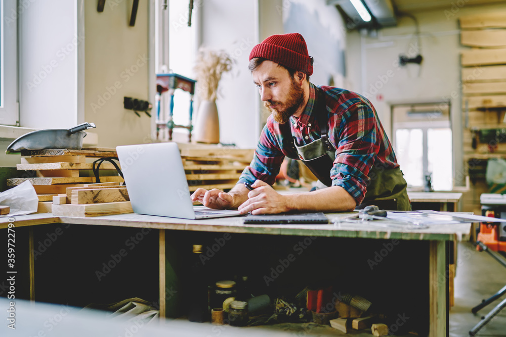Young hipster guy watching training tutorial on laptop computer learning at carpentry courses, skilled male artisan using netbook for searching information during woodworks and making plannings. - obrazy, fototapety, plakaty 