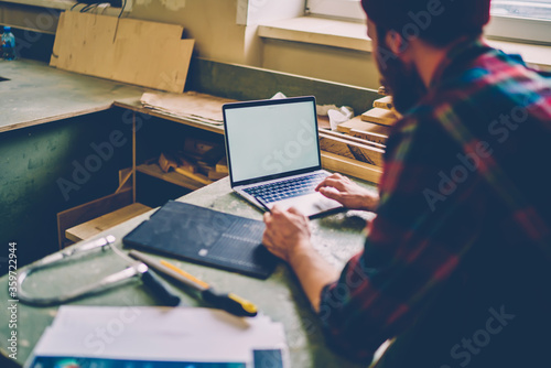 Anonymous repairman in casual shirt chatting on laptop at workshop