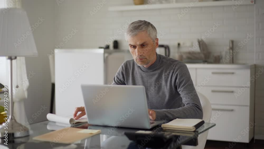 Tracking zoom in medium shot of senior man with grey hair working on laptop computer, closing pc and thinking sitting at table at home Tracking zoom in medium shot of senior man with grey hair working on laptop computer, closing pc and thinking sitting at table at home