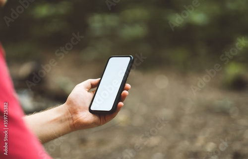 Fototapeta Naklejka Na Ścianę i Meble -  A man holds a smartphone with a white screen in a forest clearing while hiking.