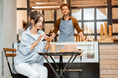 Young female customer sitting with phone at the cafe or pastry shop with a seller on the background