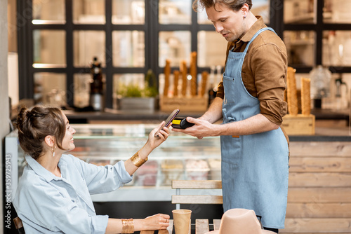 Happy waiter with a client at the cafe or ice-cream shop, making contactless payment with a smartphone