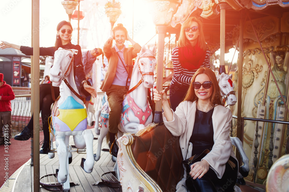 Group of attractive young women having fun riding on carousel in amusement park, girlfriends enjoying time together while riding on a merry go round during vacation holidays