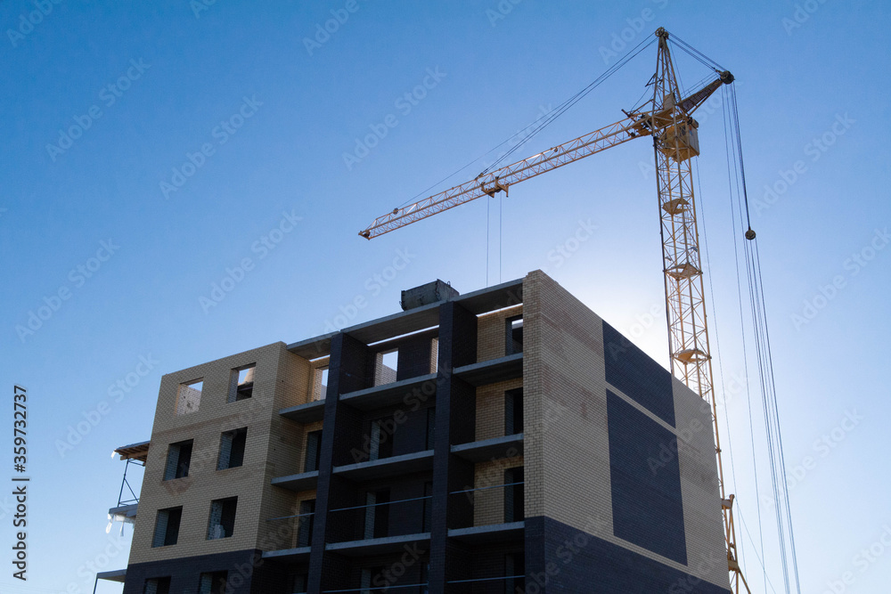 Brick building under construction and a high-rise crane against a blue sky. Construction of an office, house. A shot against the sun for an advertising company in real estate industry and business.