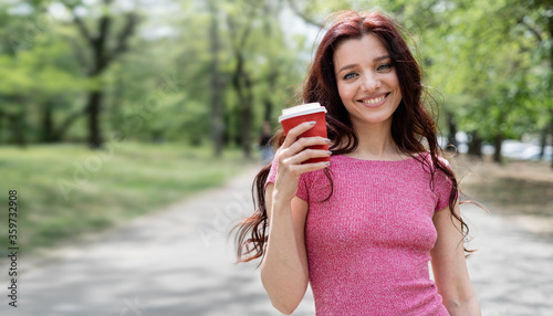 A model girl with dark hair walks in the park and drinks takeaway coffee in a glass. A slender and fashionable young lady 