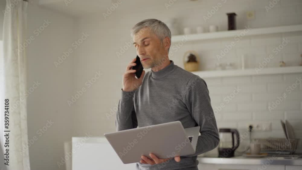 Senior man talking on cell phone pacing back and forth at home with laptop in his hands. Businessman finishing call, sitting down at desk, working on pc and thinking Senior man talking on cell phone pacing back and forth at home with laptop in his hands. Businessman finishing call, sitting down at desk, working on pc and thinking