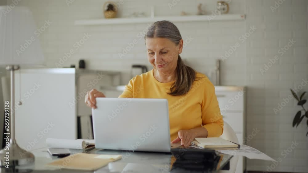 Tracking medium shot of senior woman working on laptop computer, closing pc, looking at camera and smiling sitting at table in domestic kitchen Tracking medium shot of senior woman working on laptop computer, closing pc, looking at camera and smiling sitting at table in domestic kitchen