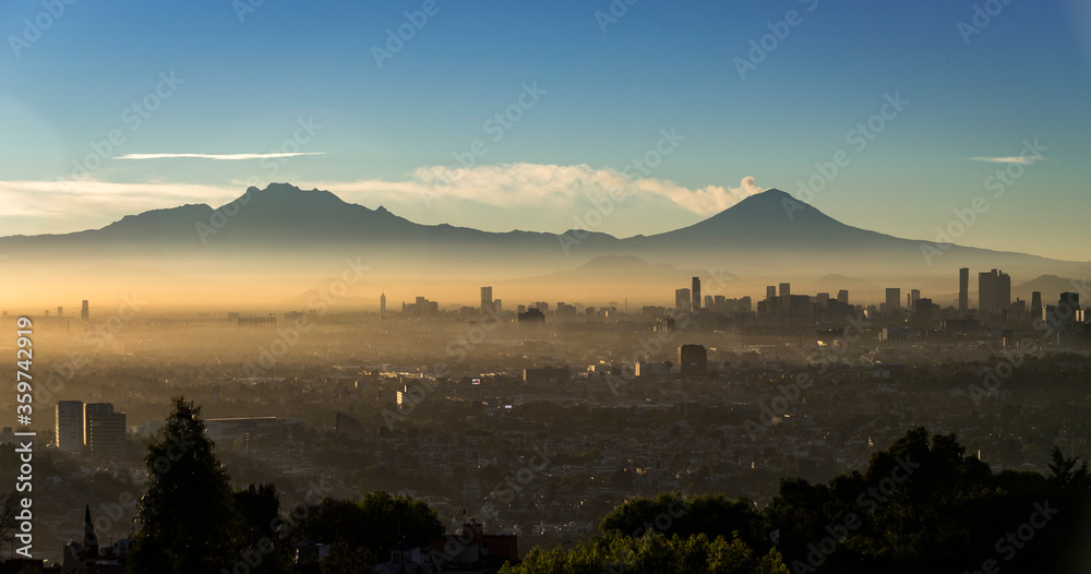 Obraz premium Panoramic view of Mexico City at dawn, with fumarole of active volcano Popocatepetl beside Iztaccihuatl, mist between skyscrapers