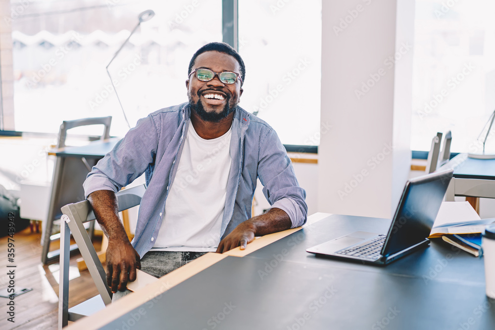 Happy casually dressed dark skinned male laughing sitting at desktop enjoying working process, cheerful african american hipster guy satisfied with online business spending time in coworking space. - obrazy, fototapety, plakaty 