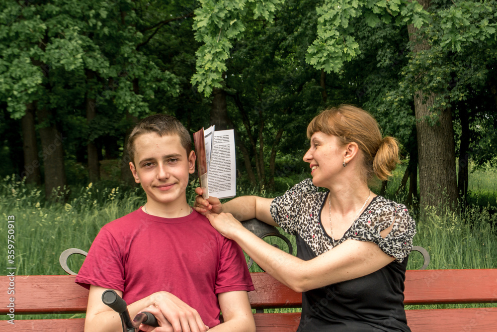 Fototapeta premium Family vacation in the park on a bench. Mom and son spend time outdoors reading an interesting book. Pleasant and useful - rest and enlightenment. Dialogue of generations..