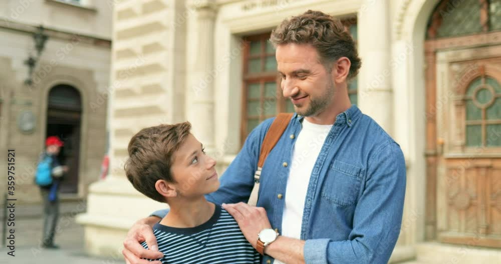 Portrait shot of Caucasian young cheerful happy man standing at street in city with his cute little son. Teen boy with dad hugging and smiling to camera outdoor. Portrait shot of Caucasian young cheerful happy man standing at street in city with his cute little son. Teen boy with dad hugging and smiling to camera outdoor.