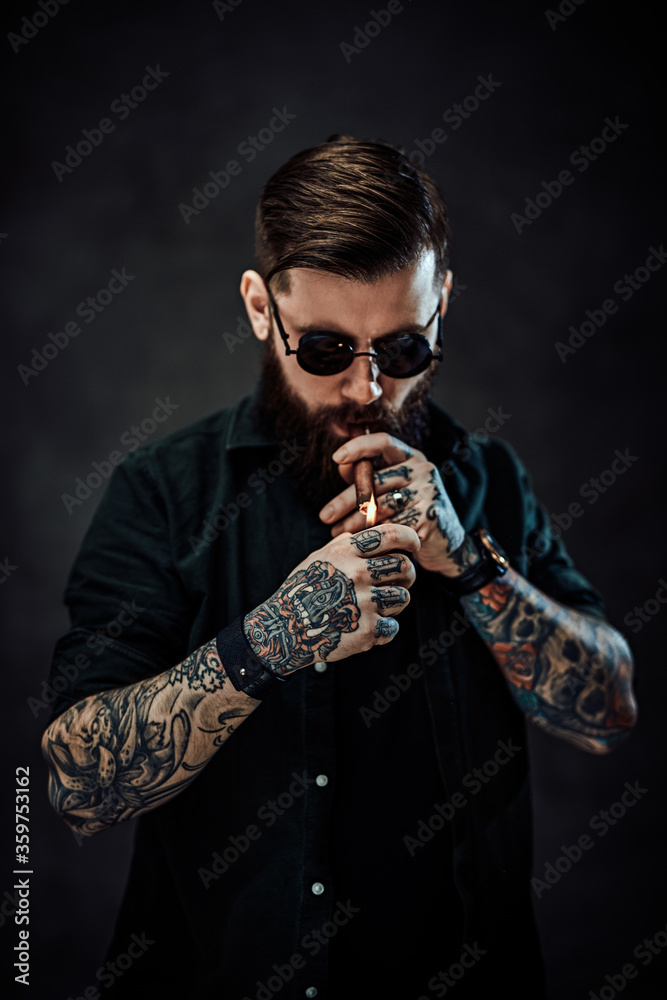 Naklejka premium Portrait of a bearded tattooed guy wearing green shirt and sunglasses lights a cigar