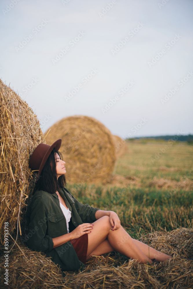 Stylish girl relaxing at hay bale in summer field in sunset. Young woman in hat sitting at haystack, atmospheric tranquil moment. Countryside slow life Stylish girl relaxing at hay bale in summer field in sunset. Young woman in hat sitting at haystack, atmospheric tranquil moment. Countryside slow life