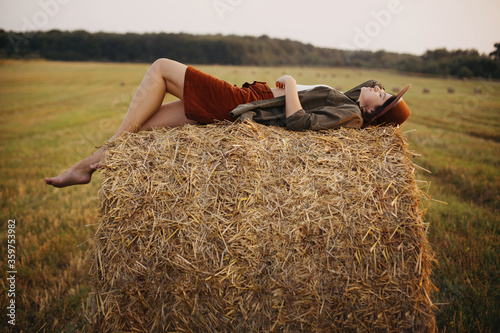 Stylish girl relaxing on hay bale in summer field in sunset. Young woman in hat resting on hay in sunshine, atmospheric tranquil moment. Countryside slow life